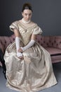 A Victorian woman wearing a gold evening gown and sitting on a pink, antique sofa