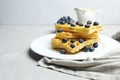 Viennese or Belgian fresh waffles on a white plate with blueberries. In the background is a bowl of berries and creamer. Morning