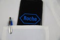 Close up of mobile phone screen with logo lettering of Roche pharmaceutical company in pocket of white doctors coat with pencil