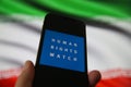 View on smartphone screen with logo lettering of human rights watch organization. Blurred Iran flag background.