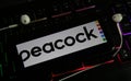 Closeup of smartphone screen with logo lettering of online tv and movie streaming service peacock on illuminated computer keyboard