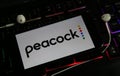 Closeup of smartphone screen with logo lettering of online tv and movie streaming service peacock on illuminated computer keyboard