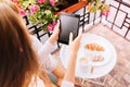 View from above tablet in hands of girl in pajama sitting surround flowers on balcony, having breakfast in the morning.