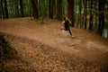 View from above of young woman in black sportswear jogging downhill on forest trail, tall leafy trees on background