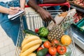 View of african american man pointing with finger at shopping cart with groceries near woman with notebook