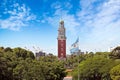 View Of The Argentinean Flag And A Red Clock Tower Behing Green Trees, Against A Blue Summer Sky With A Few White Clouds