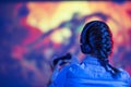 View from back of gamer girl playing video game at home in front of big screen with joystick and headphone. Colorful neon led