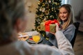 View from back of loving grandmother giving festive box with Christmas present to cheerful granddaughter sitting at Royalty Free Stock Photo