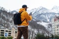 View from back. Man tourist in yellow hoodie, cap with backpack stands on background of high snowy mountains and using smartphone Royalty Free Stock Photo