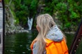 View from the back to the head of a beautiful young blonde girl in an orange life jacket standing on the edge and looking far in a