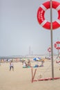 View of a beach with lifeguard lookout post, with lifebuoy and surfboard. Beach with people sunbathing and seawall, lighthouse as