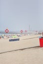 View of a beach with lifeguard lookout post, with lifebuoy and surfboard. Beach with people sunbathing and seawall, lighthouse as