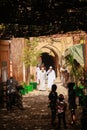 View beyond shade on two muslim men in white robes walking in sunshine alleyway, bright arches background, blurred playing kids