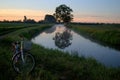 View of a bicycle on the edge of a country road and a tree reflected in the water surface of the canal in the morning sun. Royalty Free Stock Photo