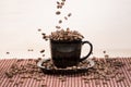 View of black cup standing on black plate with falling down roasted coffee beans on tablemat