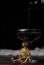 View of bottle pouring black Halloween cocktail into glass with skeleton on table and black background Royalty Free Stock Photo