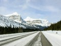 A view from a car driving down an empty highway in the winter with a huge snowy mountain in the backing along icefields parkway
