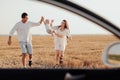 View Through Car Window, Mom and Dad Holding Their Toddler Daughter by Hands While Walking on the Field at Sunset Royalty Free Stock Photo
