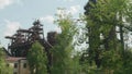 View of chimneys of old abandoned metallurgic plant through green branches of ural trees. Backlit.