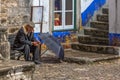 View of elderly with typical coat, sitting on wooden bench, with rain hat open on the ground, in the square of the medieval Royalty Free Stock Photo