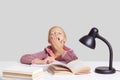 View of fatigue pretty schoolgirl covers mouth with palm, yawns as wants to sleep, being tired of studying, sits at white table, r Royalty Free Stock Photo