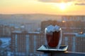 A view of a glass mug with hot black coffee and marshmallows against the backdrop of the rising sun and city skyline