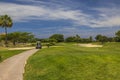 View of golf course with moving electric golf cart against backdrop of blue sky with white clouds.