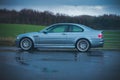 View of a gray BMW M3 CSL parked on the wet asphalt before a field