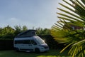 View of a gray camper van with a pop-up roof parked on the lot of a campground with sun starts and palm trees