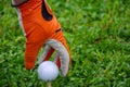 View of hand with orange glove and white golf ball on tee, on green grass, horizontal Royalty Free Stock Photo