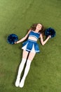 View of happy cheerleader girl in blue uniform lying with pompoms on green field