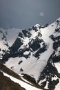 View of a high steep rocky mountain partially covered with snow against a dark cloudy sky with clouds clinging to the Royalty Free Stock Photo