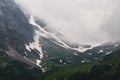 View of a high steep rocky mountain partially covered with snow against a dark cloudy sky with clouds clinging to the Royalty Free Stock Photo