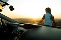 View from inside of a young woman standing near her car enjoying warm sunset view. Girl traveler leaning on vehicle hood looking Royalty Free Stock Photo