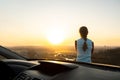 View from inside of a young woman standing near her car enjoying warm sunset view. Girl traveler leaning on vehicle hood looking Royalty Free Stock Photo