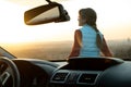 View from inside of a young woman standing near her car enjoying warm sunset view. Girl traveler leaning on vehicle hood looking Royalty Free Stock Photo