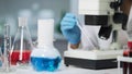 View of lab flask with evaporating blue liquid, scientist working on microscope