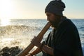 View of man sitting on seashore in sunlight and playing didjireedoo instrument. Royalty Free Stock Photo