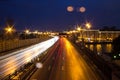 View of multilane urban highway with glowing lines of light from car headlamps and modern architecture