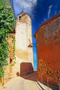 View on narrow steep empty alley uphill to medieval church bell tower, ochre fortress wall with ivy vines, blue sky - Roussillon, Royalty Free Stock Photo