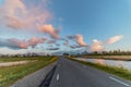 View of a paved street at sunset with grassland, spring flowers and water on both sides, with pink clouds in the air, against a br