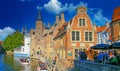 View on pier for tourist water canal boats with outside cafe terrace, medieval brick buildings against blue sky, fluffy clouds