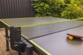 View of ping-pong racket and orange plastic ball on green netted tennis table in courtyard of villa. Royalty Free Stock Photo