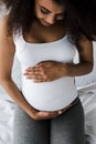 View of positive curly african american pregnant woman touching belly while sitting on bed