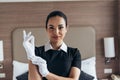 View of pretty smiling maid looking at camera and putting on white gloves in hotel room