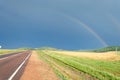 View of the road and rainbow in the sky. On the side of the road fields with green grass and forest Royalty Free Stock Photo