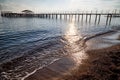 View from sand beach to water of sea, waves and pier in a nice day or evening with blues sky, bright sun and white clouds. The Royalty Free Stock Photo