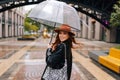 View from shoulder to positive elegant woman in fashion hat walking on city street with umbrella enjoying rainy weather Royalty Free Stock Photo