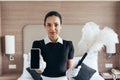View of smiling maid in white gloves holding duster and smartphone with blank screen in hotel room