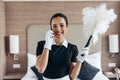 View of smiling pretty maid in white gloves holding duster and talking on smartphone in hotel room Royalty Free Stock Photo
